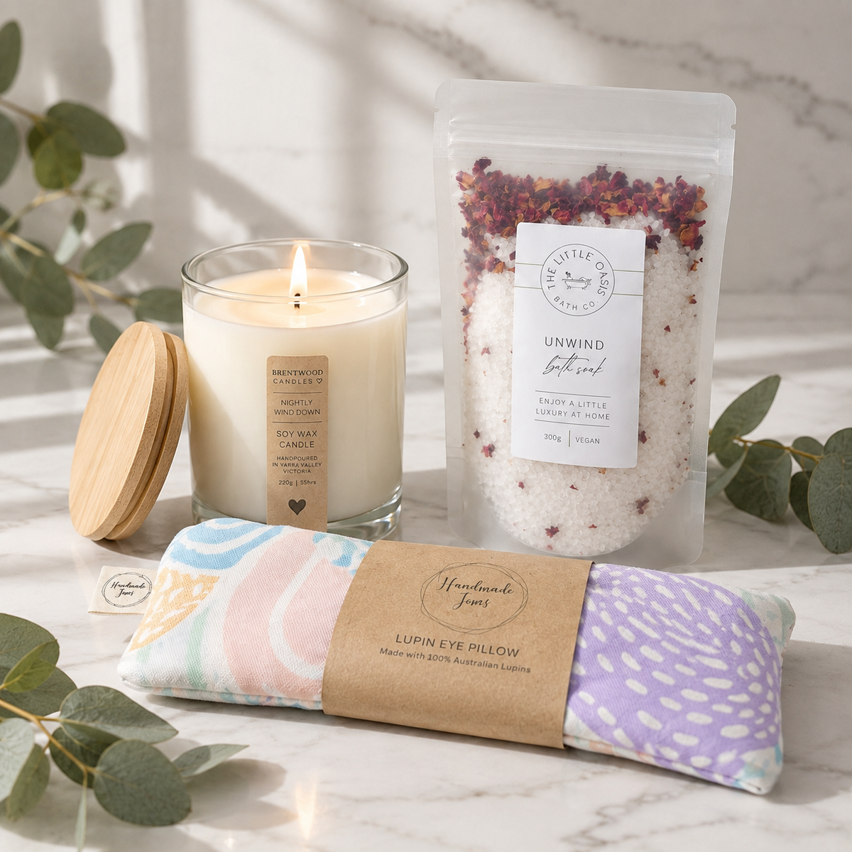 A curated self-care gift set on a light marble surface with eucalyptus leaves. The set includes a lit soy wax candle in a glass jar with a wooden lid, a stand-up pouch of bath salts, and a pastel patterned eye pillow wrapped in a kraft paper band.