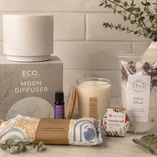 Relaxation products including a ceramic aromatherapy diffuser, candle, eye pillow, and muscle rub on a surface with a tiled wall background.