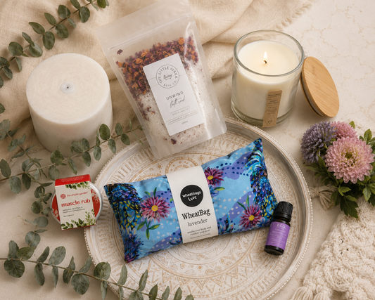 A styled flat-lay of a wellness gift kit on a cream fabric background, featuring a blue floral WheatBag, a white candle, a glass jar candle with a wooden lid, a small muscle rub packet, and an essential oil drop bottle on a white decorative tray.