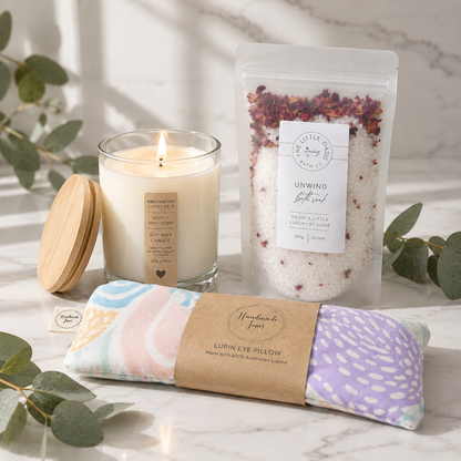A curated self-care gift set on a light marble surface with eucalyptus leaves. The set includes a lit soy wax candle in a glass jar with a wooden lid, a stand-up pouch of bath salts, and a pastel patterned eye pillow wrapped in a kraft paper band.